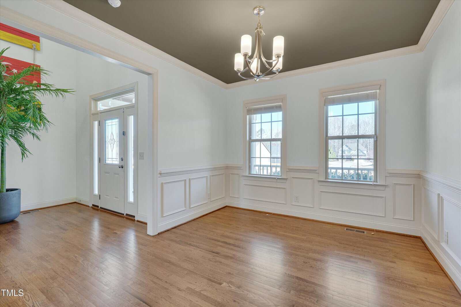 A bright, empty room with hardwood floors, white wainscoting, two windows, a chandelier, and a green ceiling. A front door with glass panels and sidelight is visible, along with part of a potted plant.