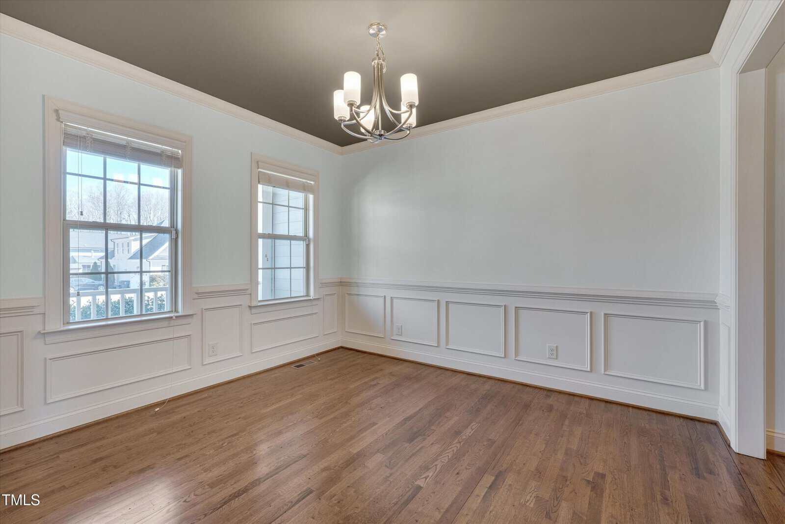 Empty dining room with light hardwood floors, white paneled walls, two large windows, a dark gray ceiling, and a modern chandelier centered on the ceiling. Natural light enters through the windows.