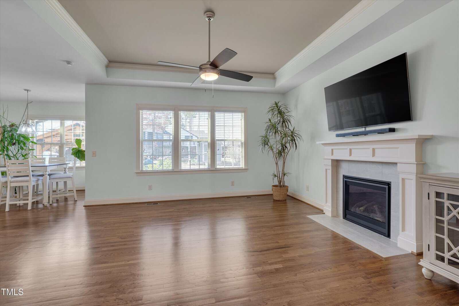 Bright living room with hardwood floors, large windows, ceiling fan, potted plant, wall-mounted TV above a fireplace, and dining area visible in the background. Room is unfurnished except for a cabinet by the fireplace.