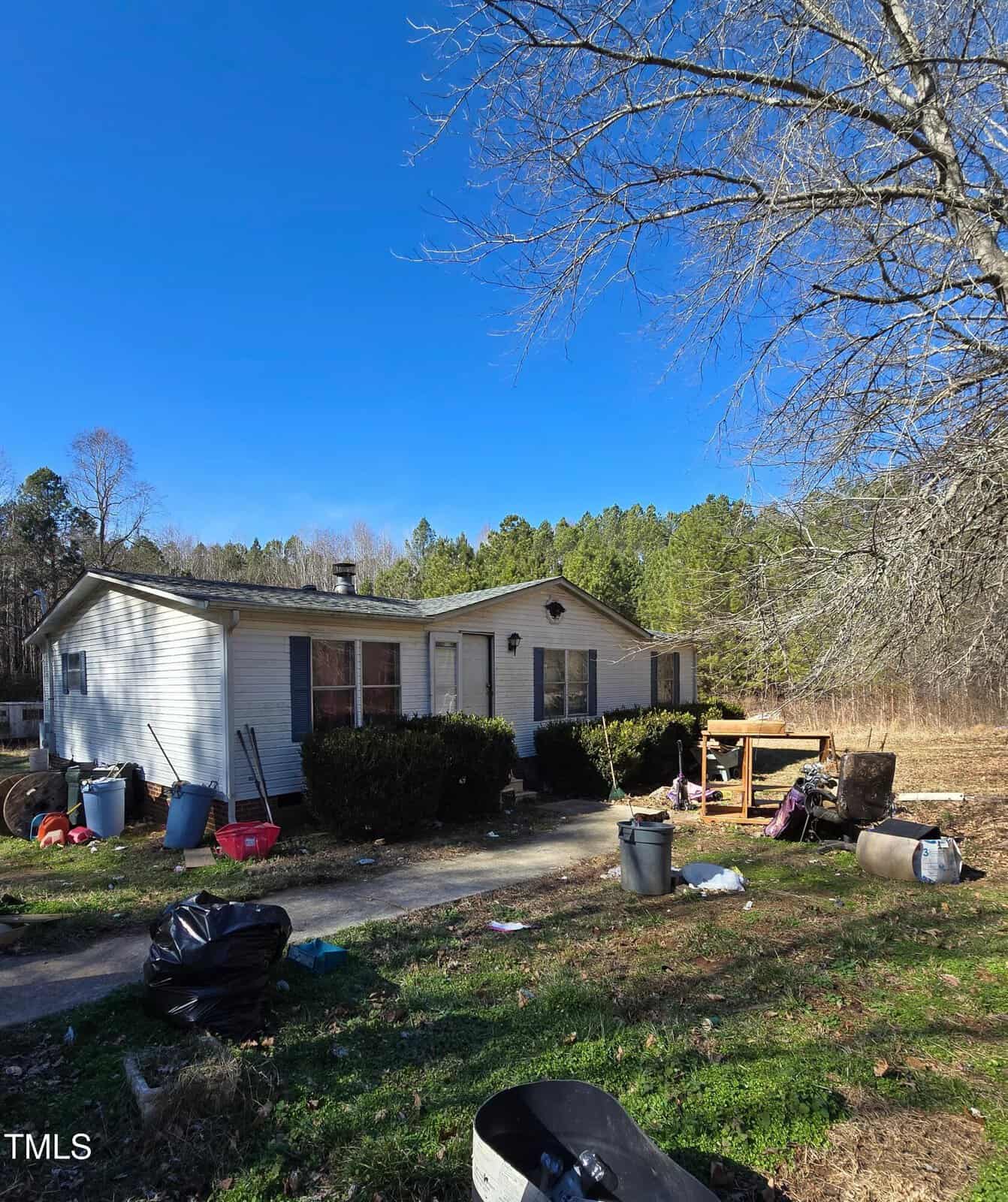 A small, worn-out house at 2383 Egypt Mountain Road with a white exterior and a shingled roof sits under a clear blue Kittrell sky. The yard is cluttered with bins, bags, and a chair. Trees and a grassy field stretch out in the background.