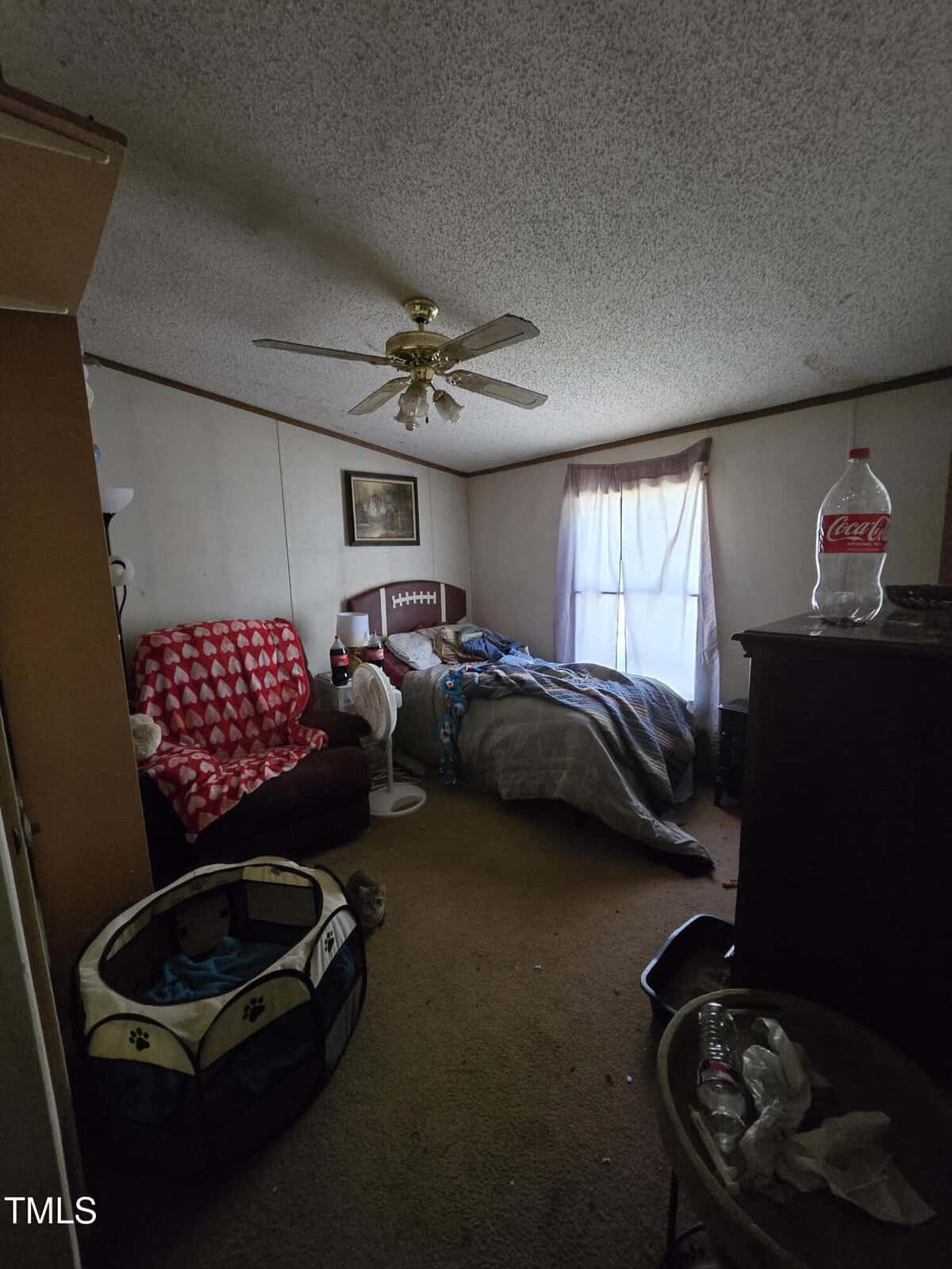 A dimly lit bedroom features a bed with rumpled covers, a ceiling fan, a red armchair with a patterned throw, a pet playpen, and a dresser with a large bottle on top. A curtain-draped window lets in some light. The floor is carpeted.