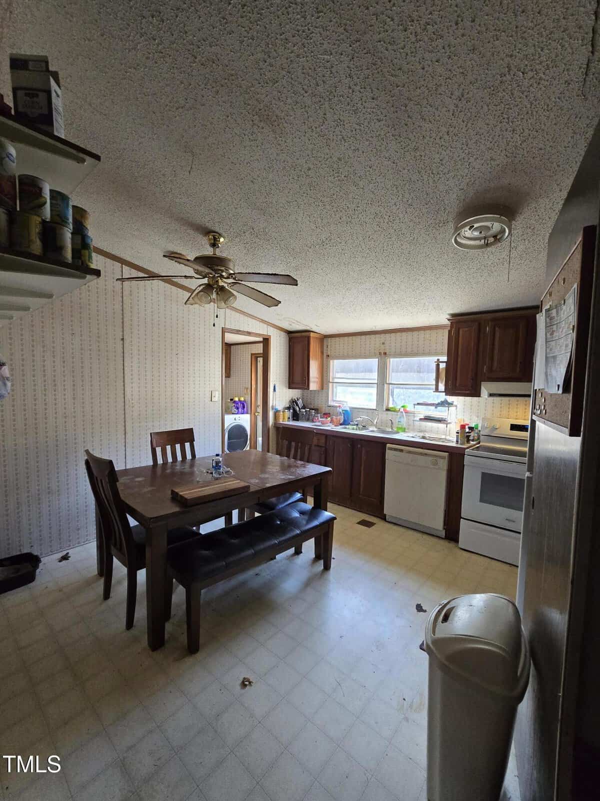 A kitchen with a wooden dining table and four chairs. The room has a patterned wallpaper, a ceiling fan, and large windows. The kitchen features dark wood cabinets, a white stove, and a sink with dishes. There are items on shelves and the floor is tiled.