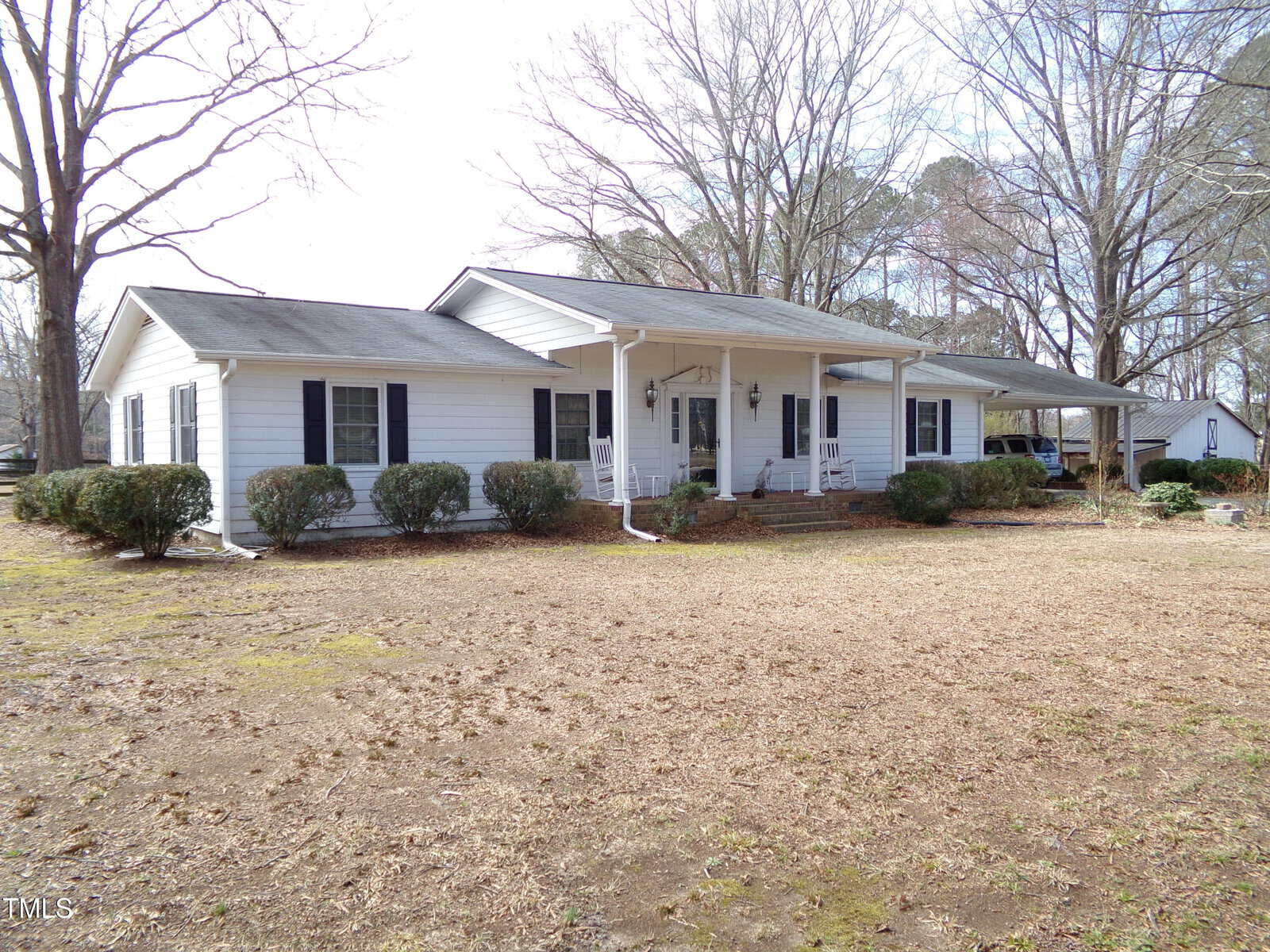 Single-story white house with a gray shingle roof, covered front porch, multiple windows, and shrubs along the front. The yard is mostly bare ground with some grass, and leafless trees are visible around the property.