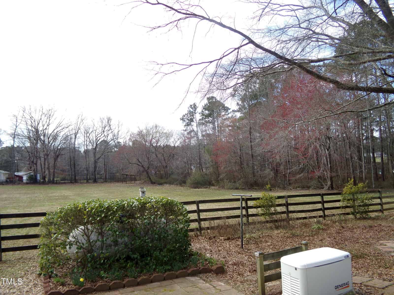 A fenced backyard with a grassy lawn, scattered trees without leaves, a bush, a white generator, and a wooden fence. The sky is overcast, and the background shows additional trees and a small farm building.
