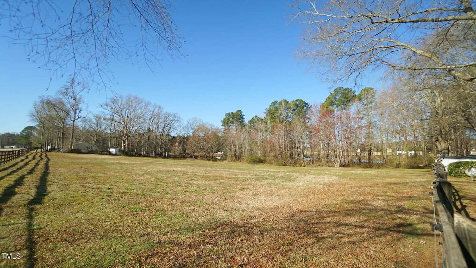 A grassy open field bordered by wooden fences with scattered trees in the background under a clear blue sky. Some areas of the trees show autumn or winter foliage. Shadows of the fence fall across the grass.