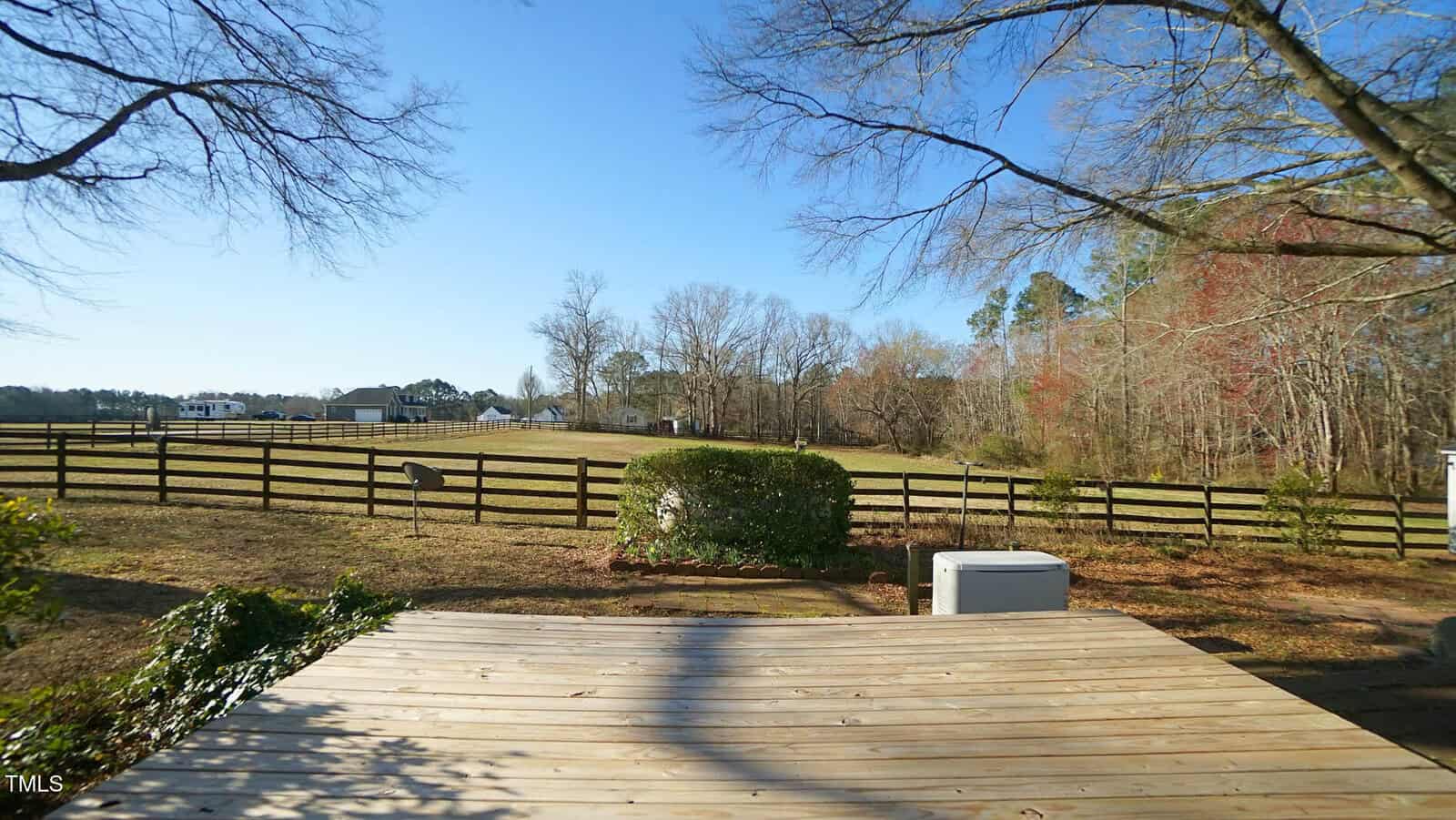 View from a wooden deck overlooking a fenced grassy backyard with trees and a clear blue sky. An HVAC unit and shrubs are near the deck, with pasture and distant buildings visible beyond the fence.