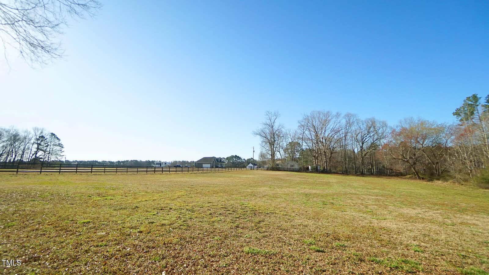 A large, open grassy field with a wooden fence on the left, trees on the right, and a few houses and farm buildings in the distance under a clear blue sky.
