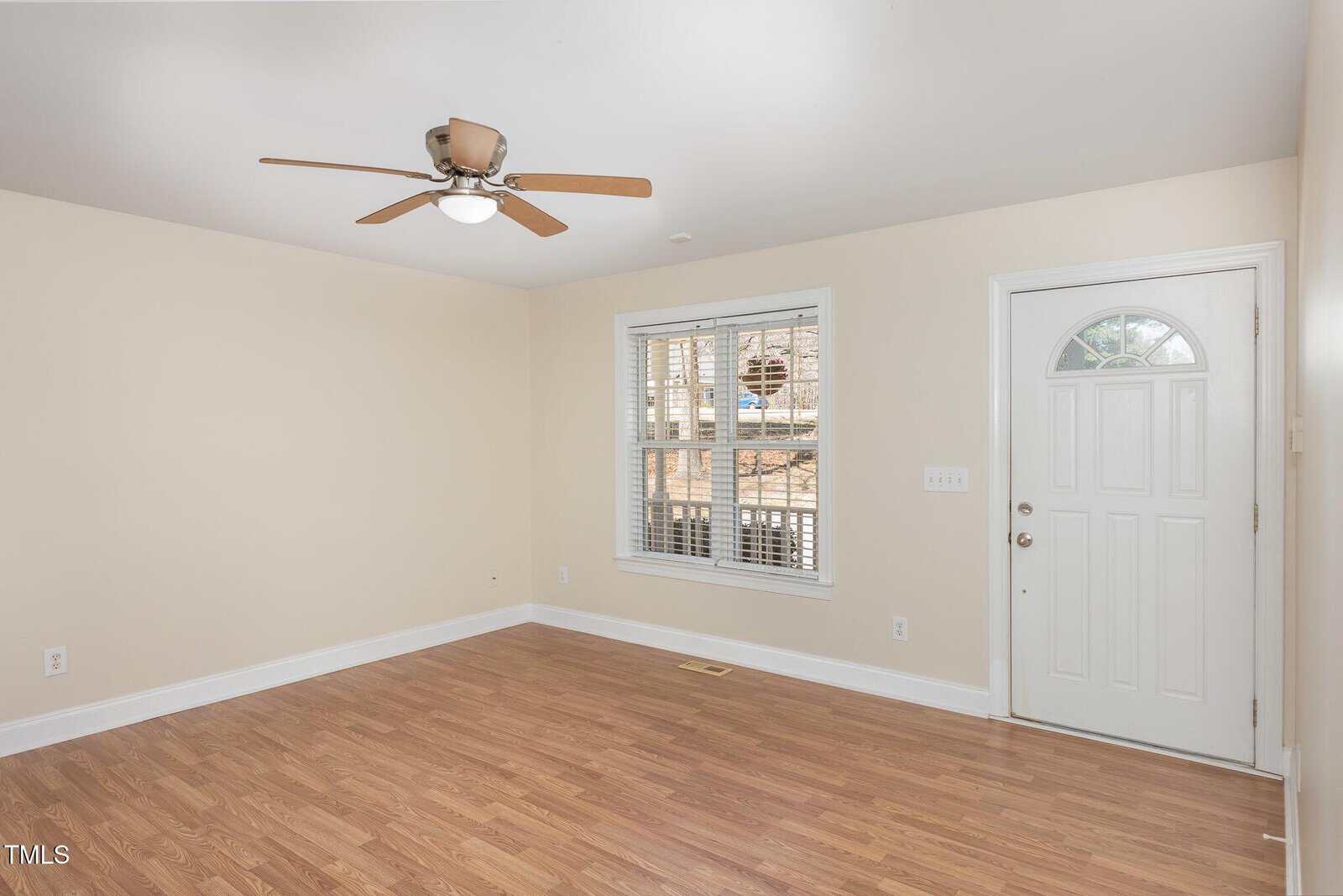 A small, empty room with beige walls, wood-look flooring, a ceiling fan with a light, a white-framed window with blinds, and a white front door with a half-circle window.