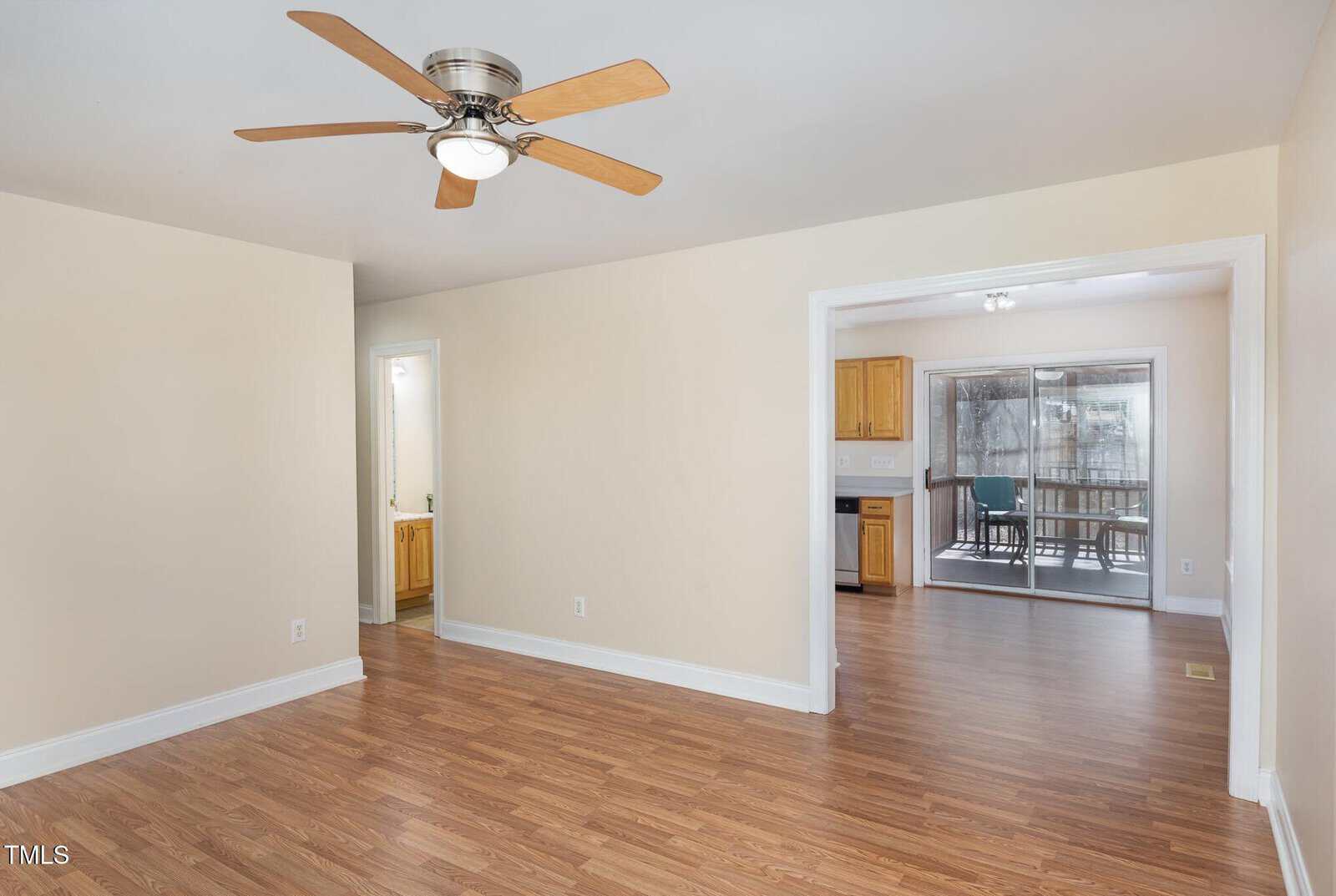 A room with light beige walls, a ceiling fan with wooden blades, and wood-look flooring. An open doorway leads to a kitchen with wooden cabinets, and a sliding glass door opens to a sunroom.