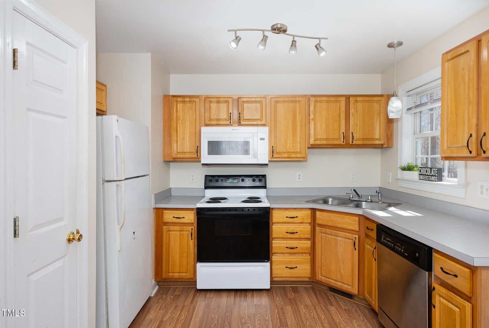 A kitchen with wood cabinets, white refrigerator, white microwave, electric stove, stainless steel dishwasher, double sink, and a window above the sink. Countertops are gray and the floor is wood.