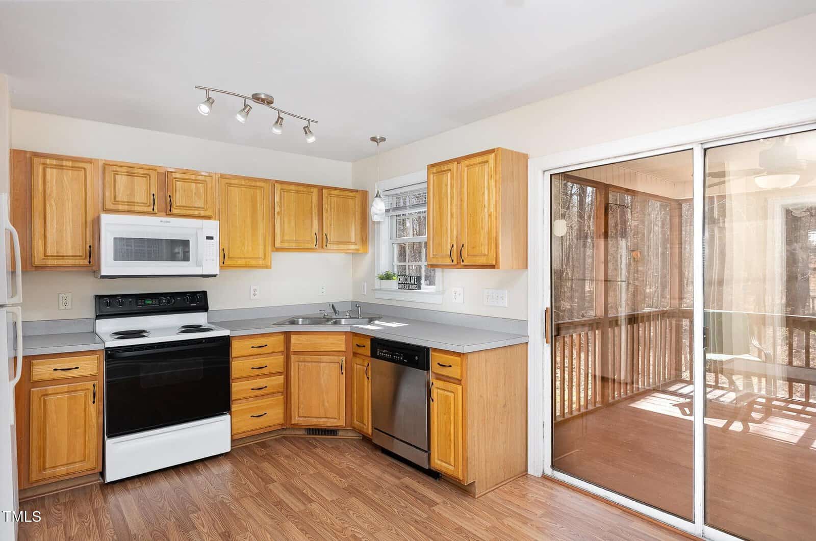 A kitchen with wooden cabinets, white countertops, black and white appliances, a stainless steel dishwasher, and a window over the sink. Sliding glass doors lead to an enclosed porch with wood floors.