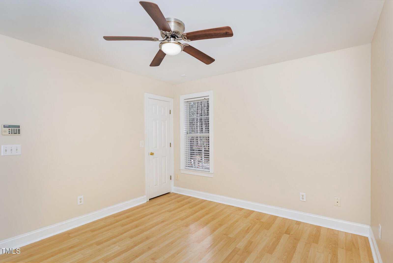 Empty room with light wood flooring, beige walls, a white door, a window with blinds, a ceiling fan with wooden blades, and several electrical outlets.