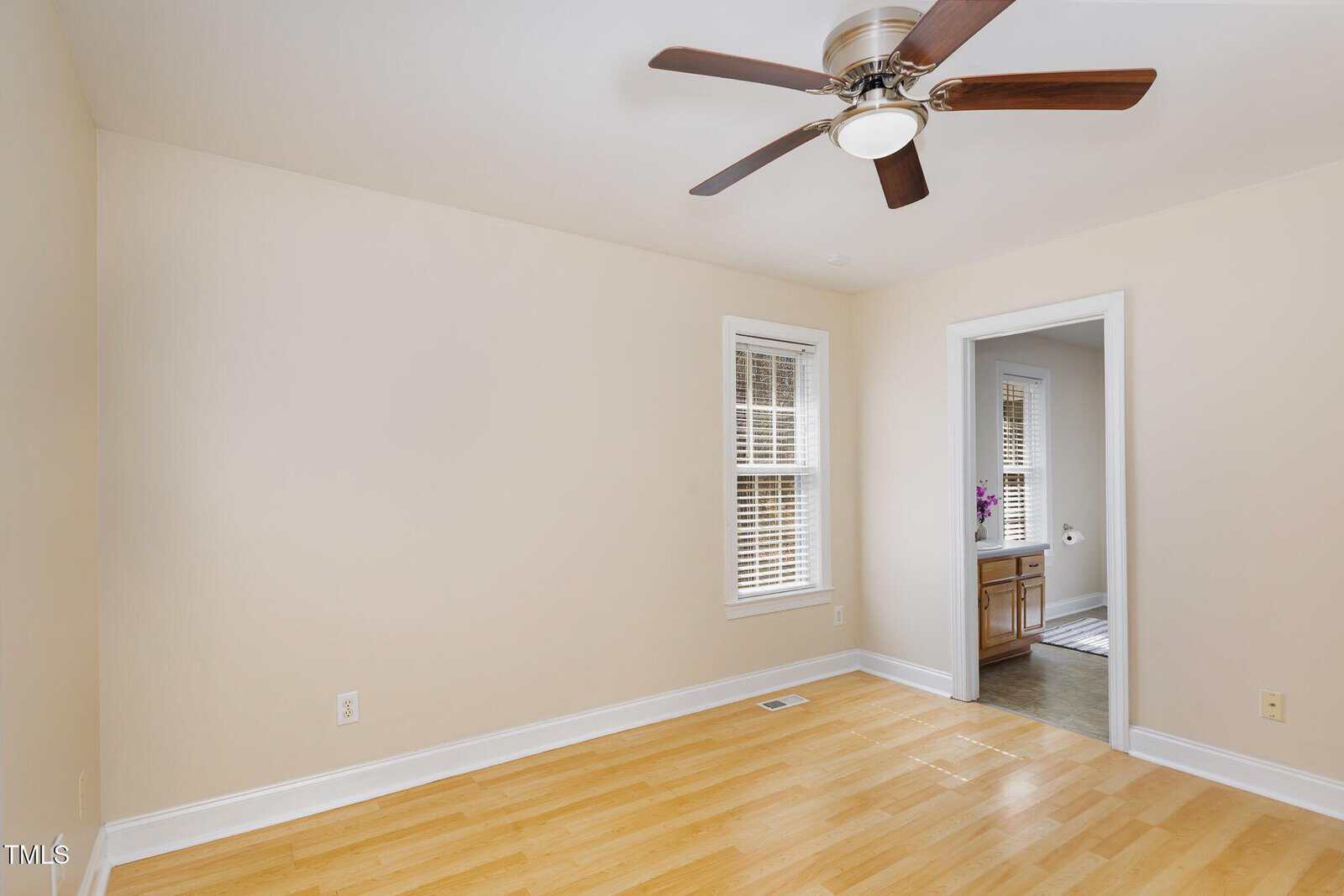 Empty room with light wood flooring, beige walls, a ceiling fan with wooden blades, two windows with blinds, and an open doorway leading to a bathroom with a visible sink and cabinet.