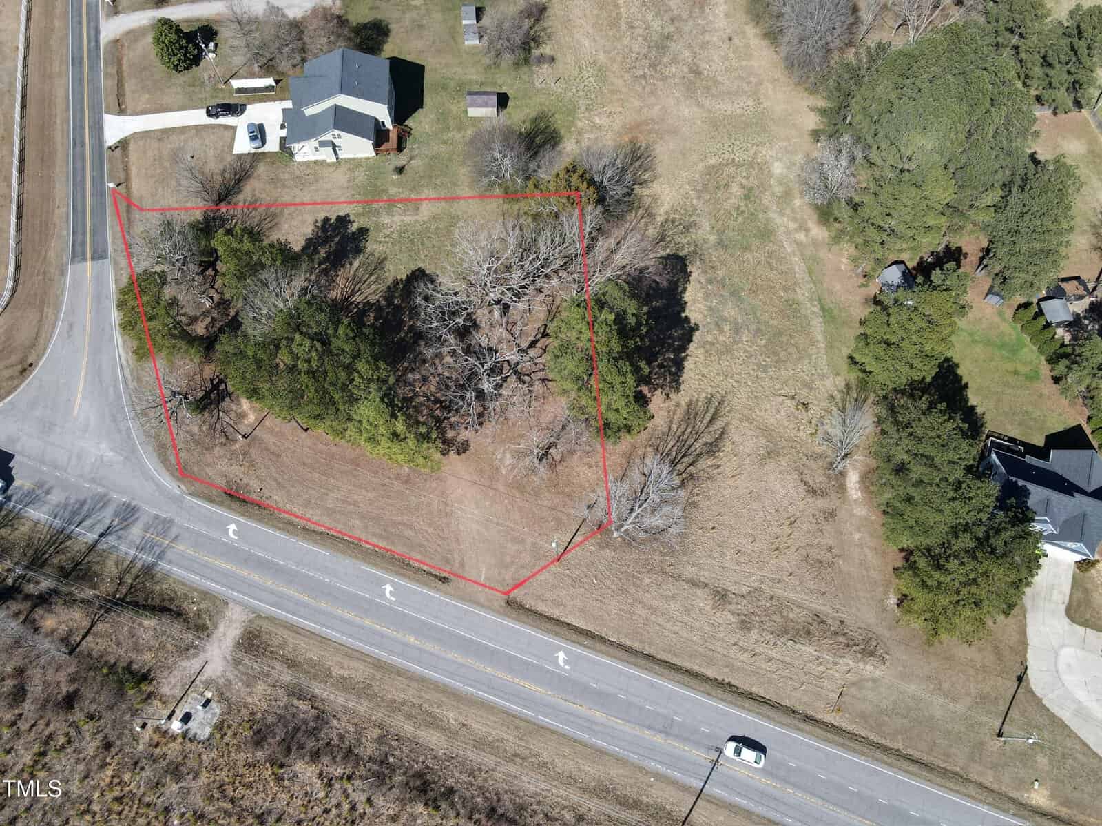 Aerial view of a vacant, partially wooded lot outlined in red at the corner of two roads, with a house and driveway on the adjacent lot; trees and open grass are visible within the outlined area.