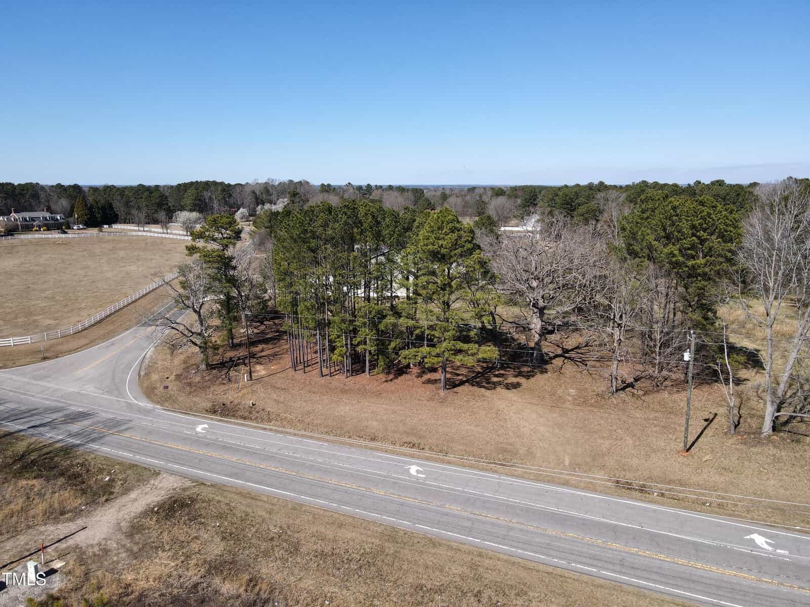 Aerial view of a rural intersection with two paved roads, bordered by grassy land and tall trees. A white fence runs along the left edge, and houses are visible in the background under a clear blue sky.