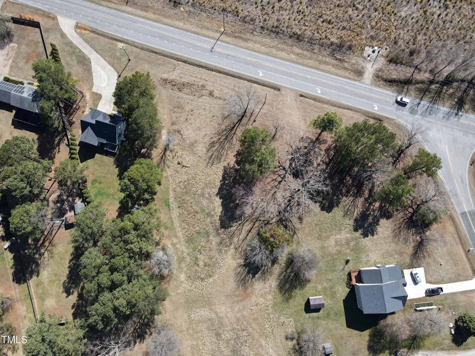 Aerial view of a grassy, partially wooded lot bordered by a road at the top and houses on either side. One house is in the bottom right and another is in the upper left, with scattered trees throughout the lot.