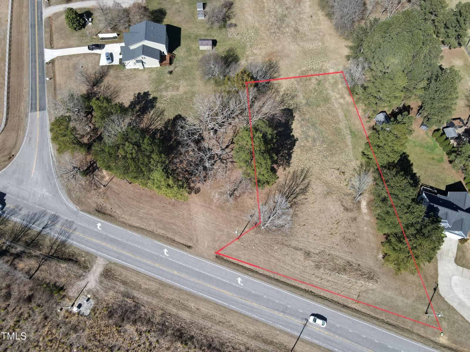 Aerial view of a grassy, empty lot outlined in red near a residential area. The lot is bordered by a paved road with painted turn arrows, trees, and several houses nearby. One car is visible on the road.