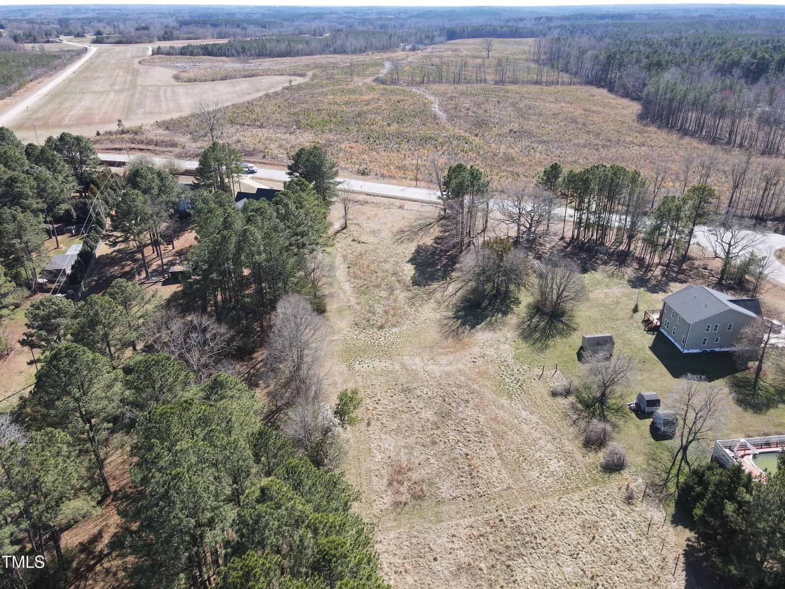 Aerial view of a rural landscape with scattered houses, trees, open grassy areas, and a road running through the middle. Fields and wooded areas extend into the distance under a clear sky.