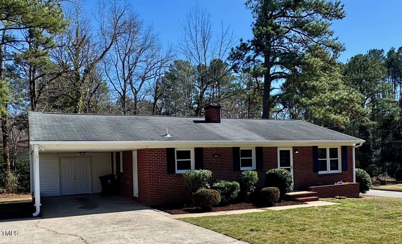One-story brick house with a dark gray shingled roof, white trim, and a single-car garage to the left. The front yard features a small lawn with neatly trimmed shrubs. Tall trees and a clear blue sky are in the background.