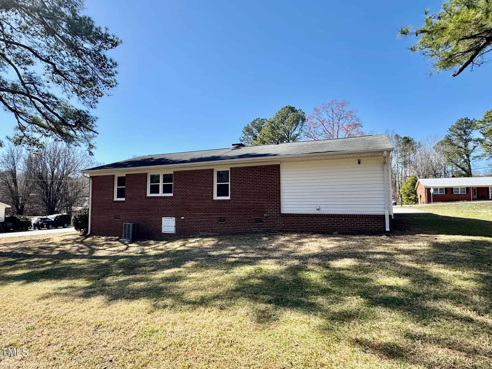 Single-story house with a red brick exterior and white siding on one side. The house has several small windows and is surrounded by a grassy lawn. Trees are visible in the background under a clear blue sky.