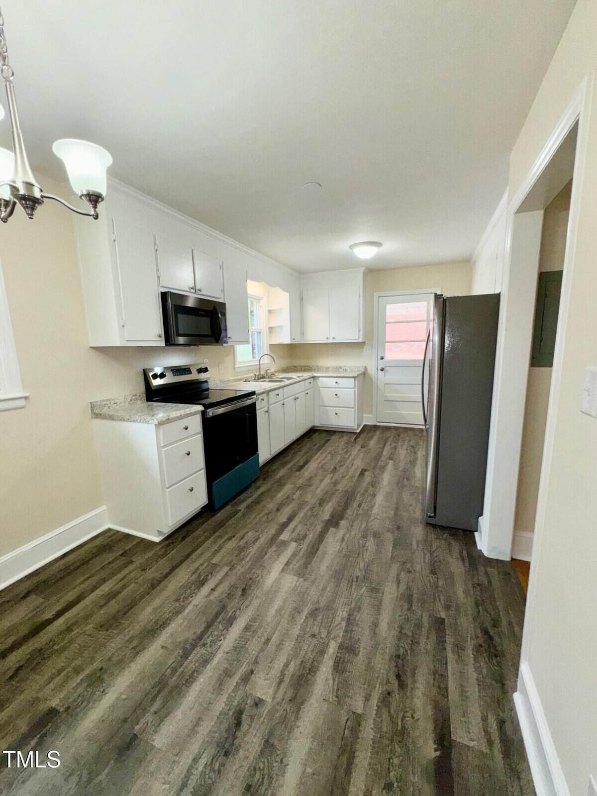 Clean kitchen with white cabinets and black hardware. Features stainless steel appliances, including a stove, microwave, and fridge. Wooden flooring and a chandelier on the left. A window and a back door are visible at the far end.