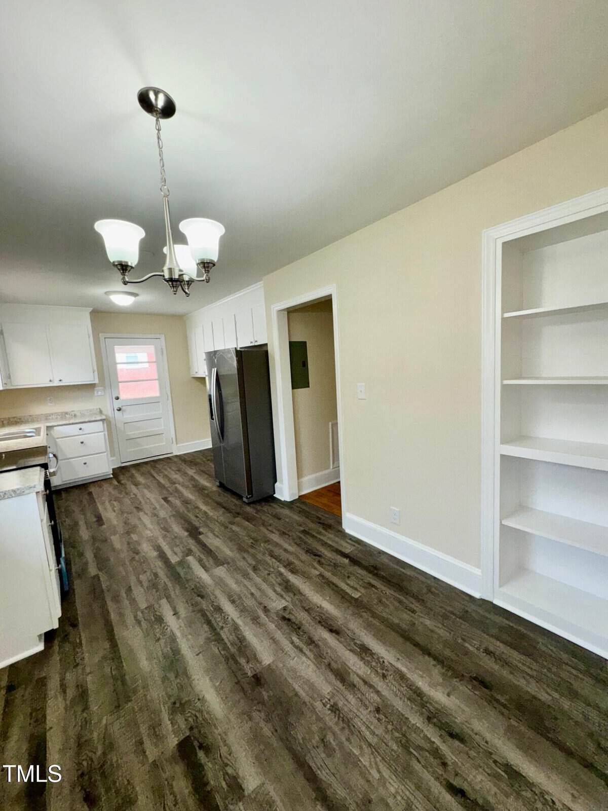 A kitchen with wood-style flooring, white cabinetry, and stainless steel appliances. A built-in bookshelf is on the right wall, and a chandelier hangs from the ceiling.