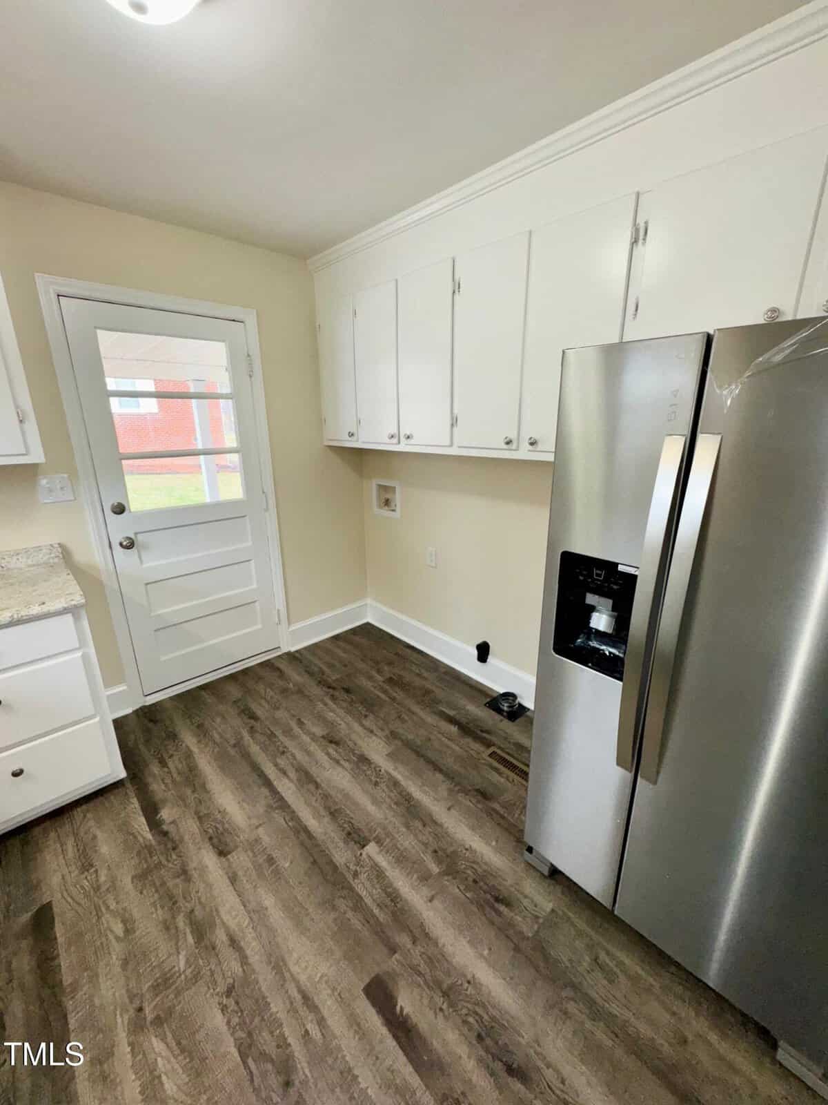 A kitchen with white cabinets, a stainless steel refrigerator, and wood-grain flooring. A small door with a window leads outside. The walls are painted light beige.