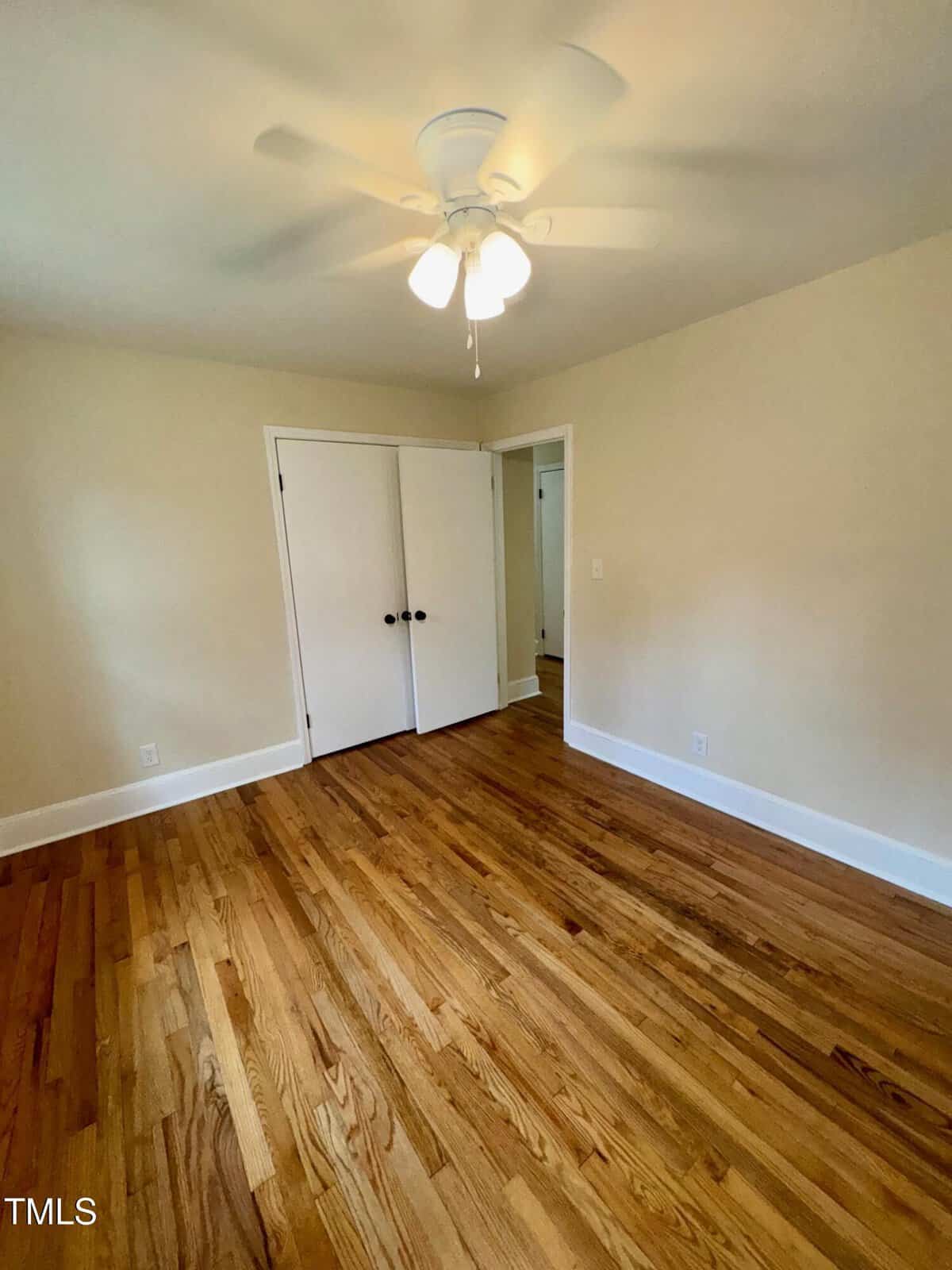 An empty room with wooden floors and beige walls. A ceiling fan with lights is mounted on the ceiling. A closed white double door is on one wall. Natural light enters from an unseen window.