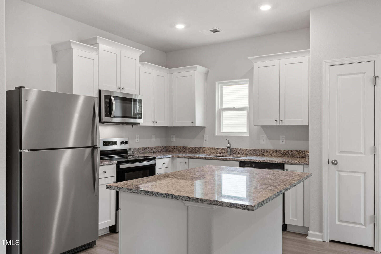 Modern kitchen with white cabinets, stainless steel appliances including a refrigerator, oven, and microwave. Features a granite-topped island with a sink. Overhead lighting and a small window provide additional illumination. White door to the right.