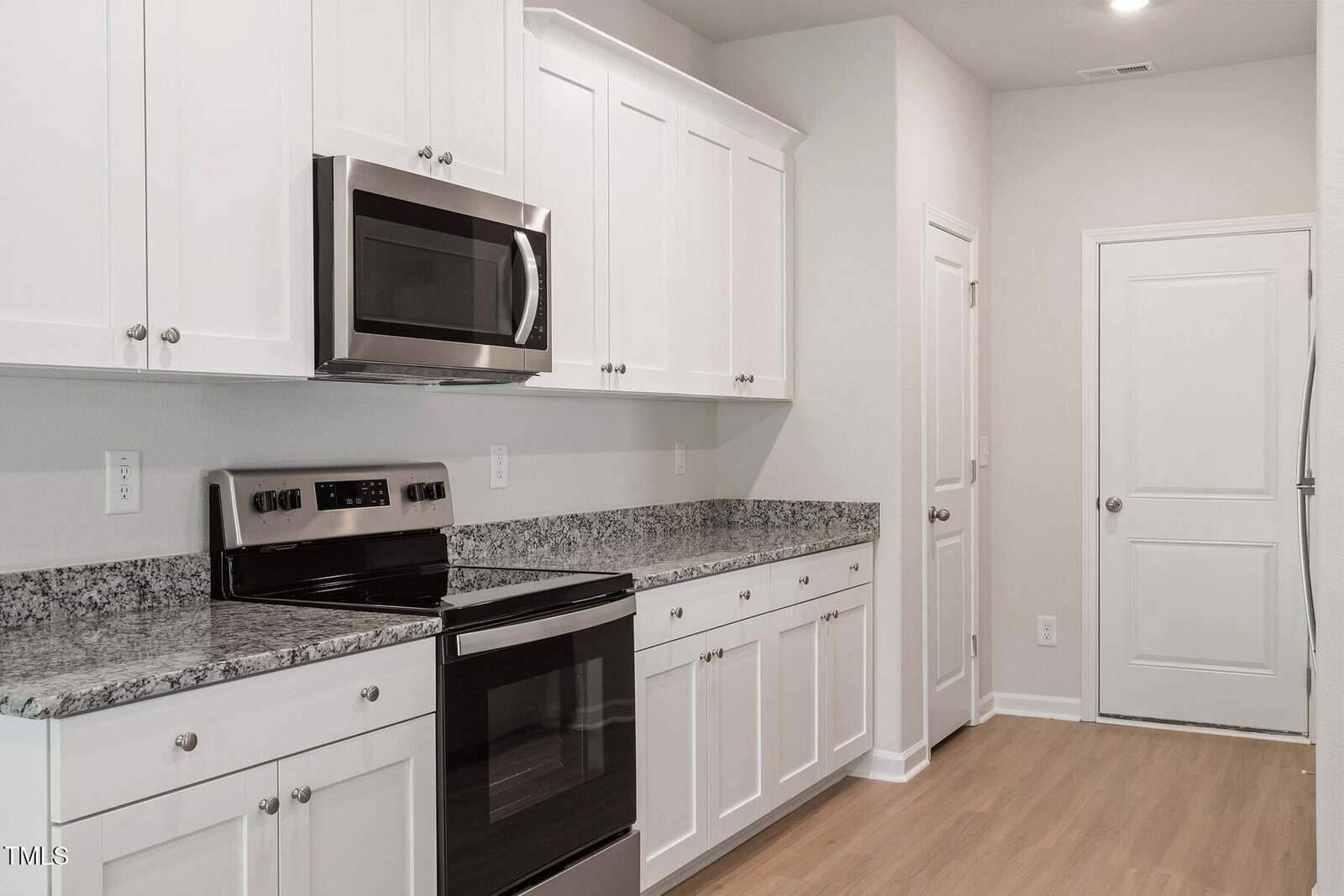 A modern kitchen with white cabinets and stainless steel appliances. There is a built-in microwave above the electric stove, granite countertops, and wooden flooring. A door is visible at the far end beside the cabinets.