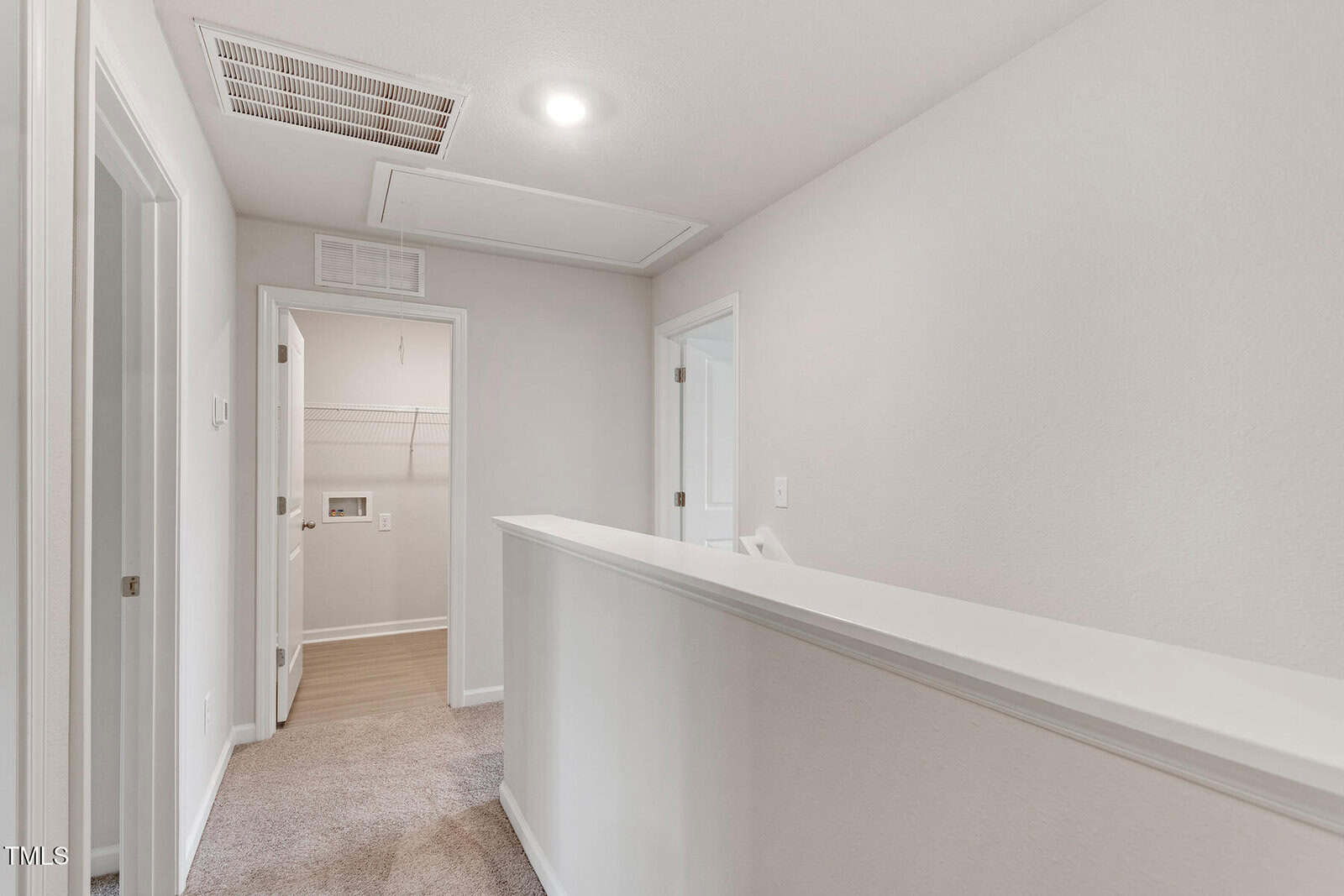 A hallway with light beige walls and carpet. The ceiling has a vent and a recessed light. There are two open doors on the left leading to a closet and laundry area. A half-wall railing is on the right.