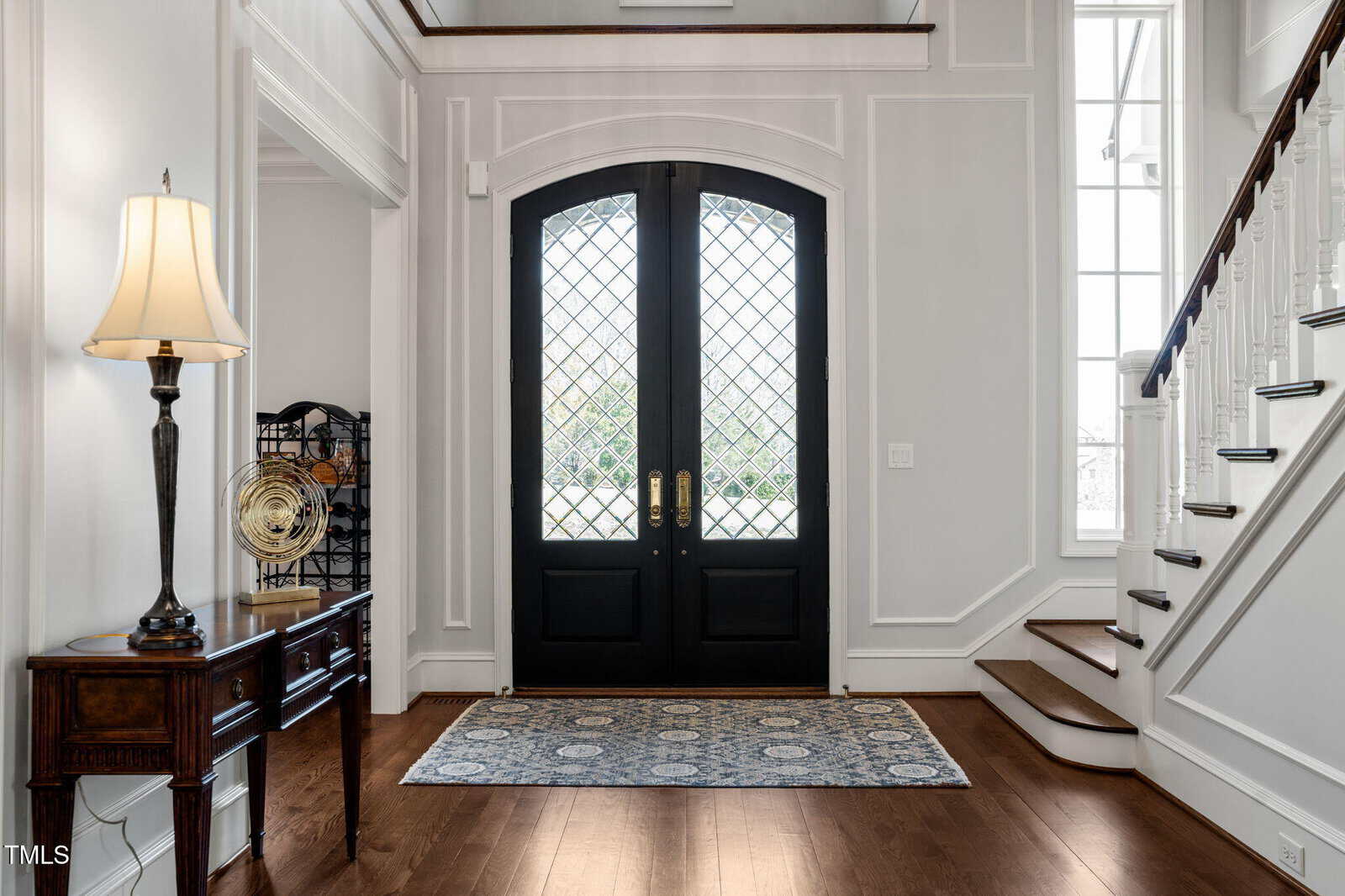 A well-lit entryway with double black doors featuring glass panes, a patterned rug, a wooden console table with a lamp, decorative items, and a staircase with white risers and wooden steps on the right.