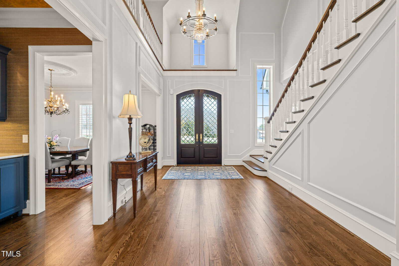 A spacious foyer with hardwood floors, a wooden staircase with white balusters, double front doors with glass panels, a chandelier, an area rug, and a table with a lamp and clock. Dining room visible to the left.