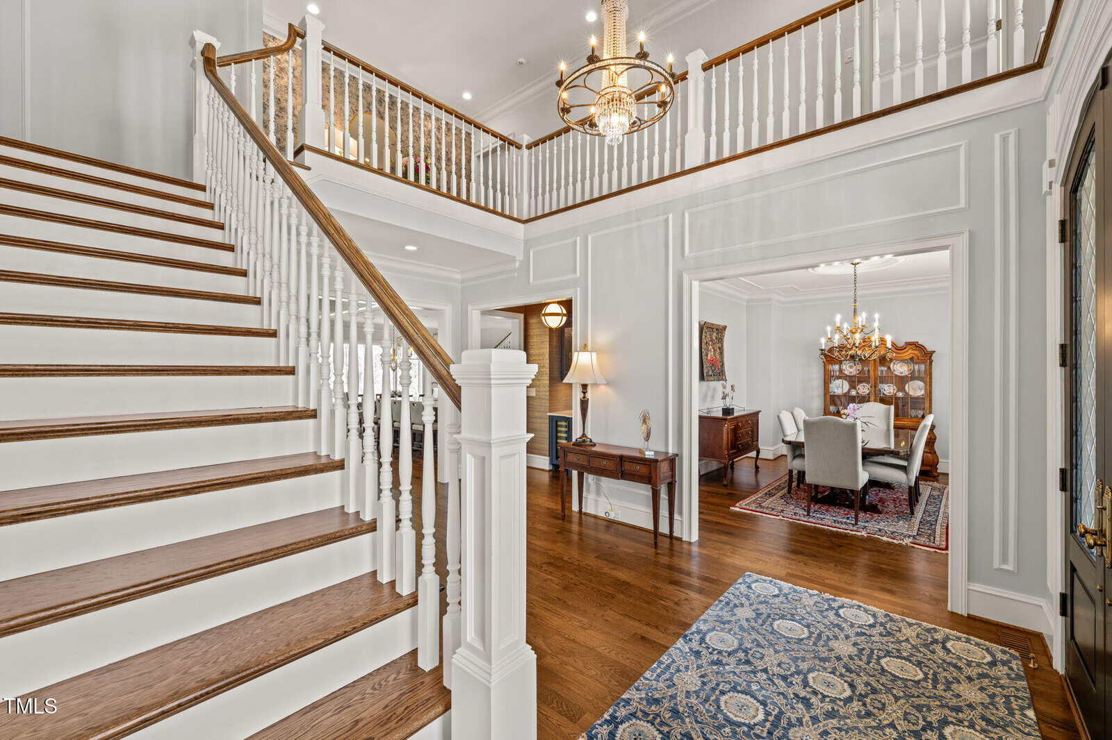 Spacious foyer with wooden staircase and white balusters, chandelier overhead, hardwood floors, and an adjacent dining room with a large wooden table and chairs visible through an open doorway.