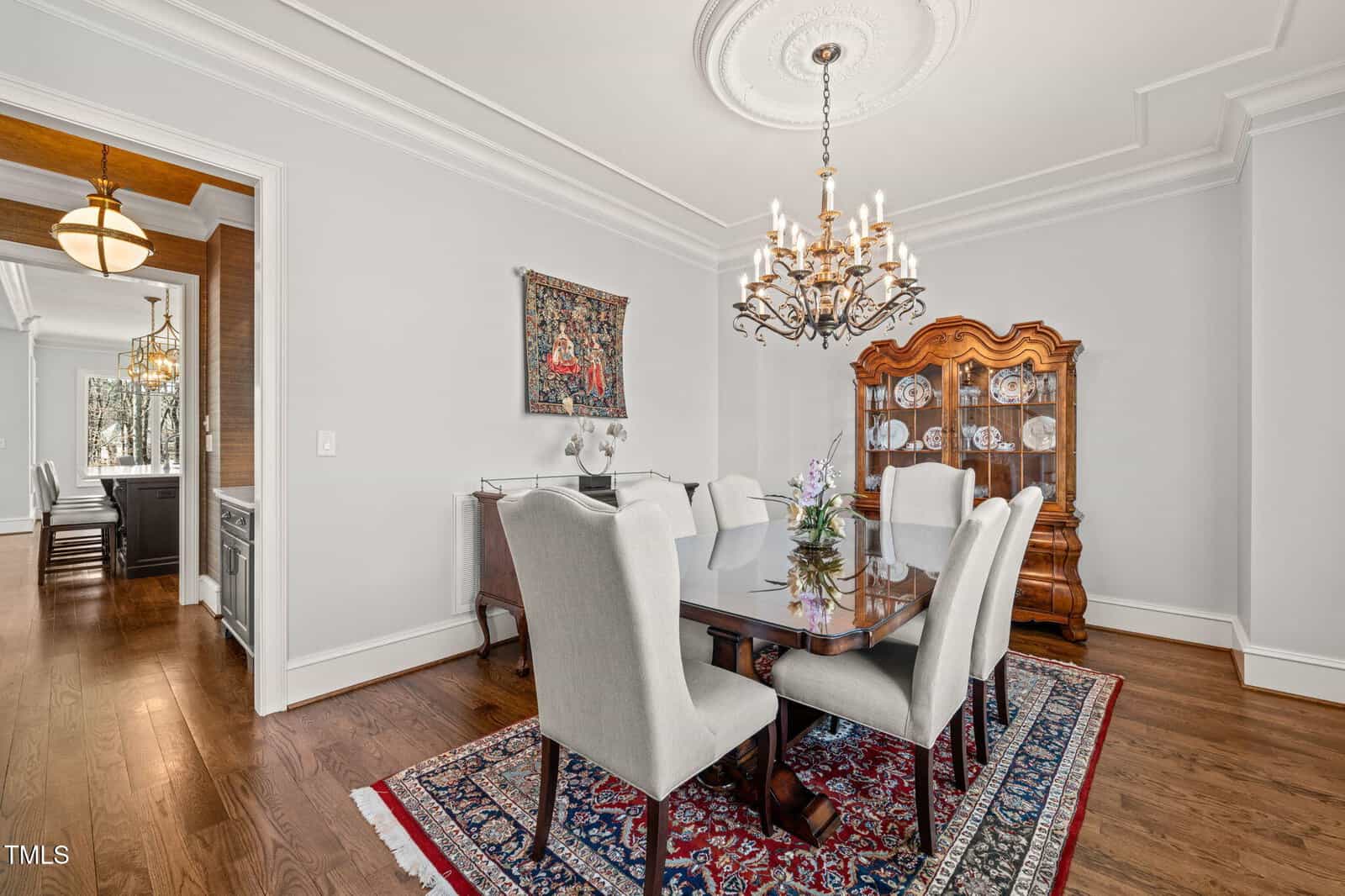 A formal dining room with a dark wood table, six upholstered chairs, a chandelier, a china cabinet, wall art, and a patterned rug on hardwood floors; adjacent room visible through an open doorway.