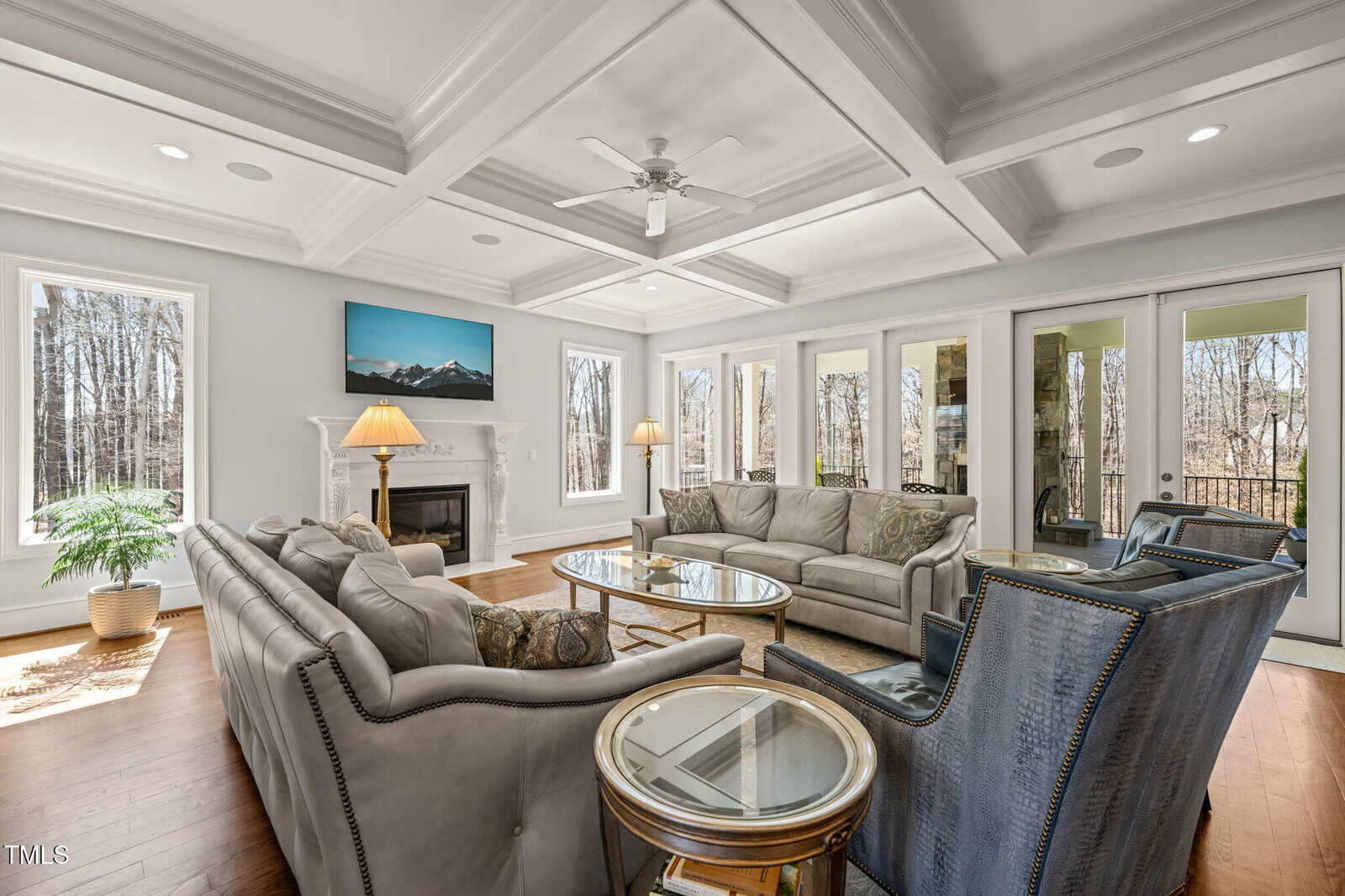 A spacious living room with coffered ceilings, large windows, two gray sofas, two blue chairs, a glass coffee table, a fireplace with a TV above it, and hardwood floors. Trees are visible outside.