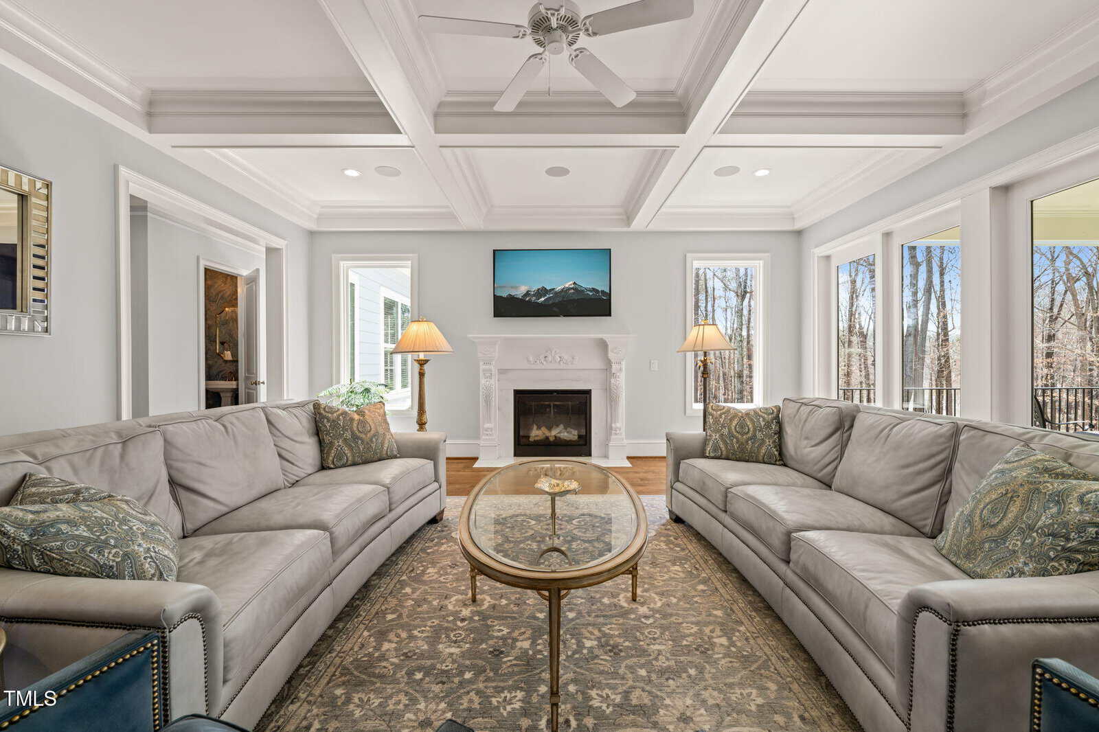 A living room with two gray sofas, a glass coffee table, an ornate rug, a fireplace with a TV above it, two lamps on either side, and large windows showing trees outside. Ceiling has a coffered design with a fan.