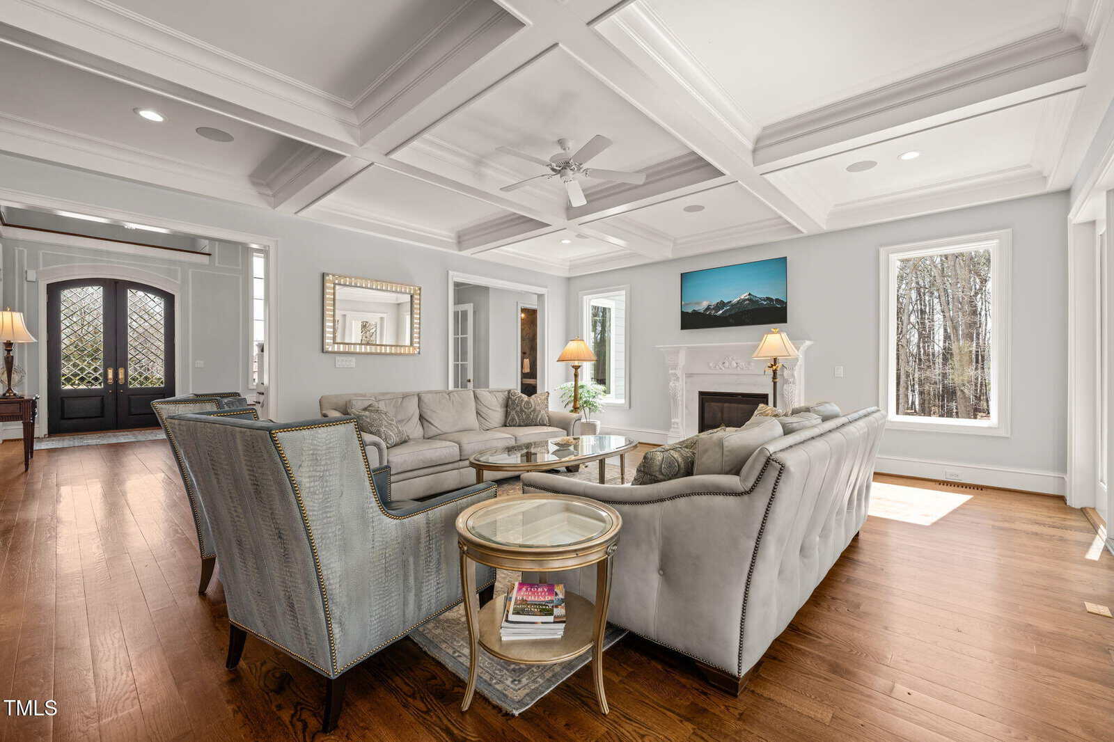 Spacious living room with two gray sofas, a matching armchair, glass-top side tables, wood floors, coffered ceiling, fireplace, wall-mounted TV, large windows, and double front doors with sidelights.