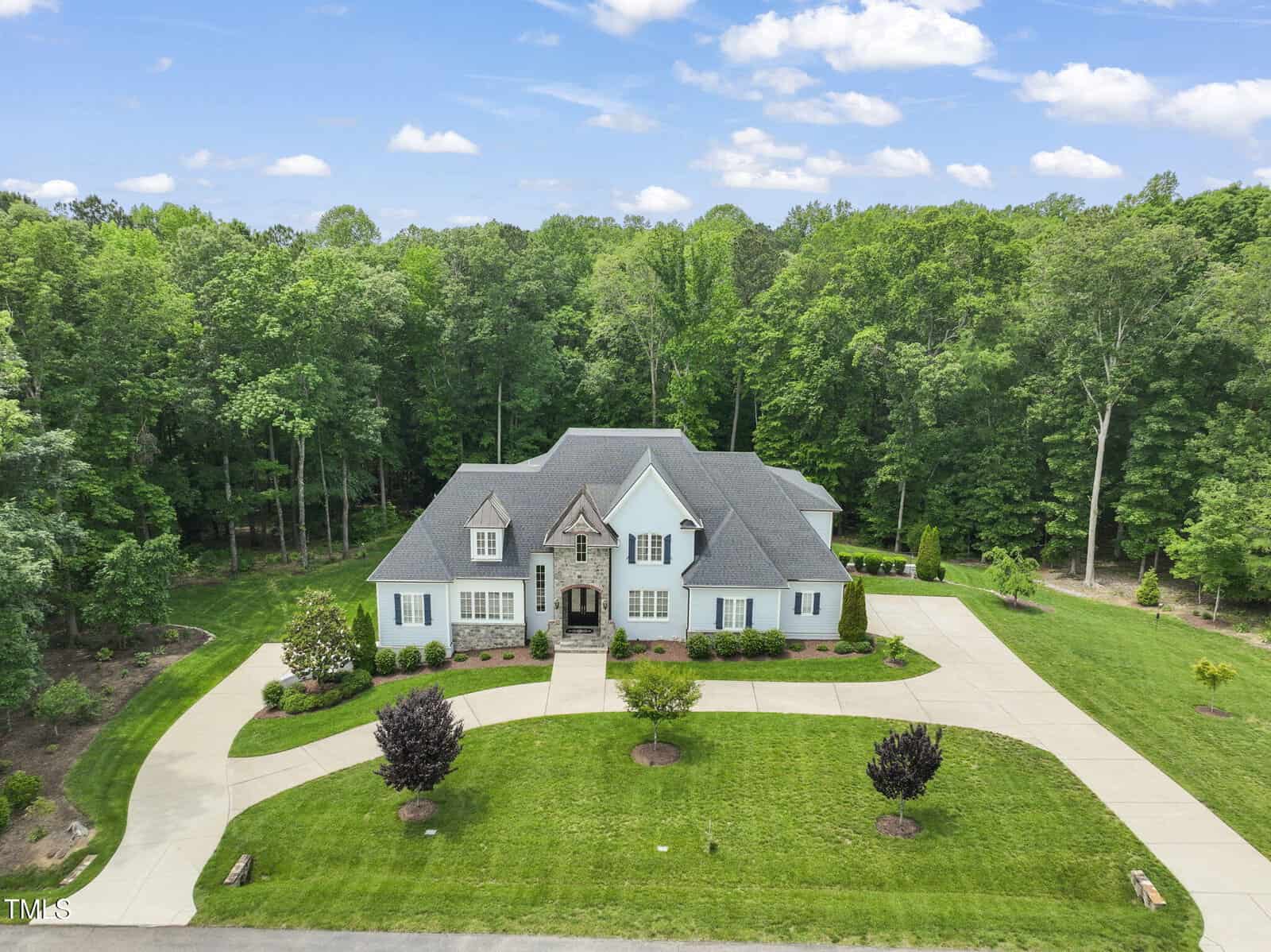A large light-colored house with dark roofing sits on a spacious, well-maintained lawn at 40 Park Meadow Lane, Youngsville NC 27596. The circular driveway is surrounded by dense trees and landscaped bushes under a partly cloudy sky.