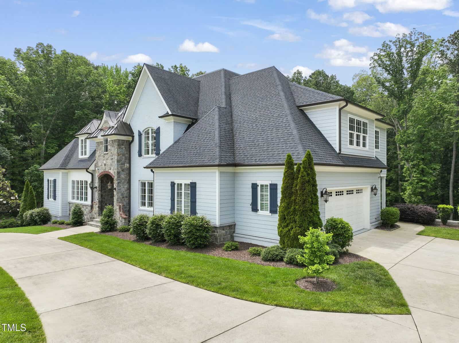 Two-story house with white siding, gray shingle roof, and stone accents. The home features multiple gables, shuttered windows, a two-car garage, and landscaped yard with shrubs and trees, all set against a wooded backdrop.