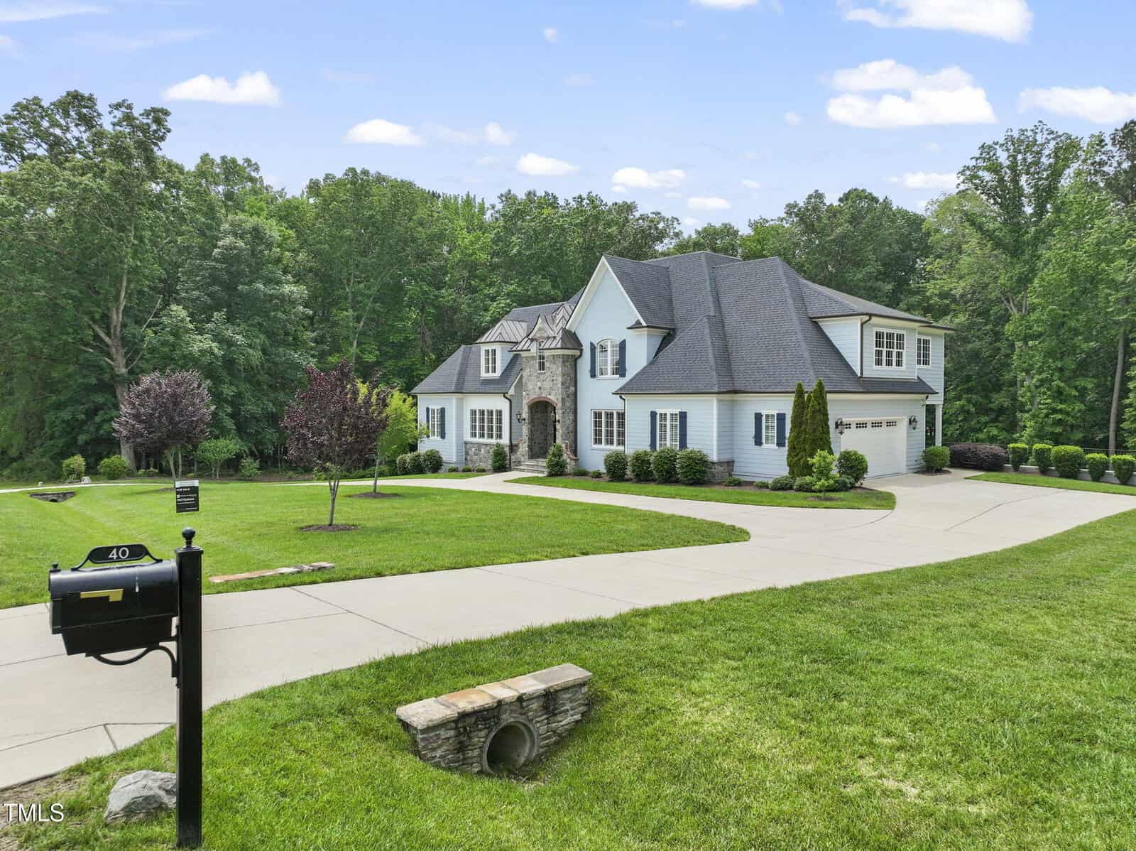 A large two-story house with light-colored siding, multiple peaks on the roof, and a three-car garage. The home is surrounded by a spacious, well-manicured lawn and trees, with a curved driveway and a black mailbox in front.