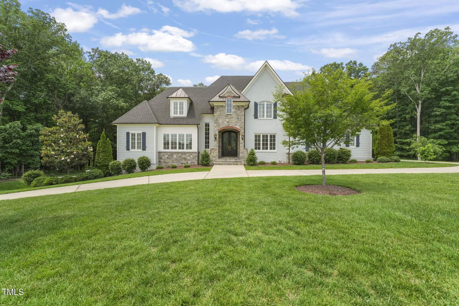 A two-story white house with stone accents, black-framed windows, and a dark front door, sits behind a curved driveway and landscaped lawn, surrounded by trees under a partly cloudy sky.