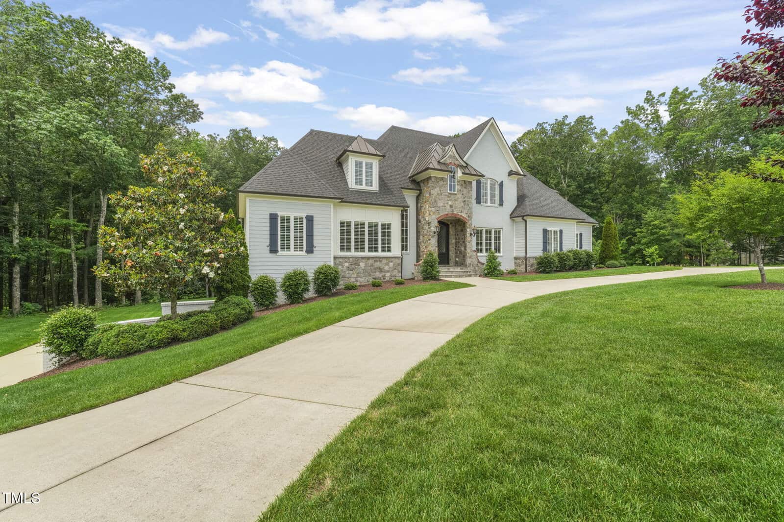 A large, two-story house with stone and white siding, multiple gables, and dormer windows sits on a well-manicured lawn with a curved concrete driveway and trees in the background.