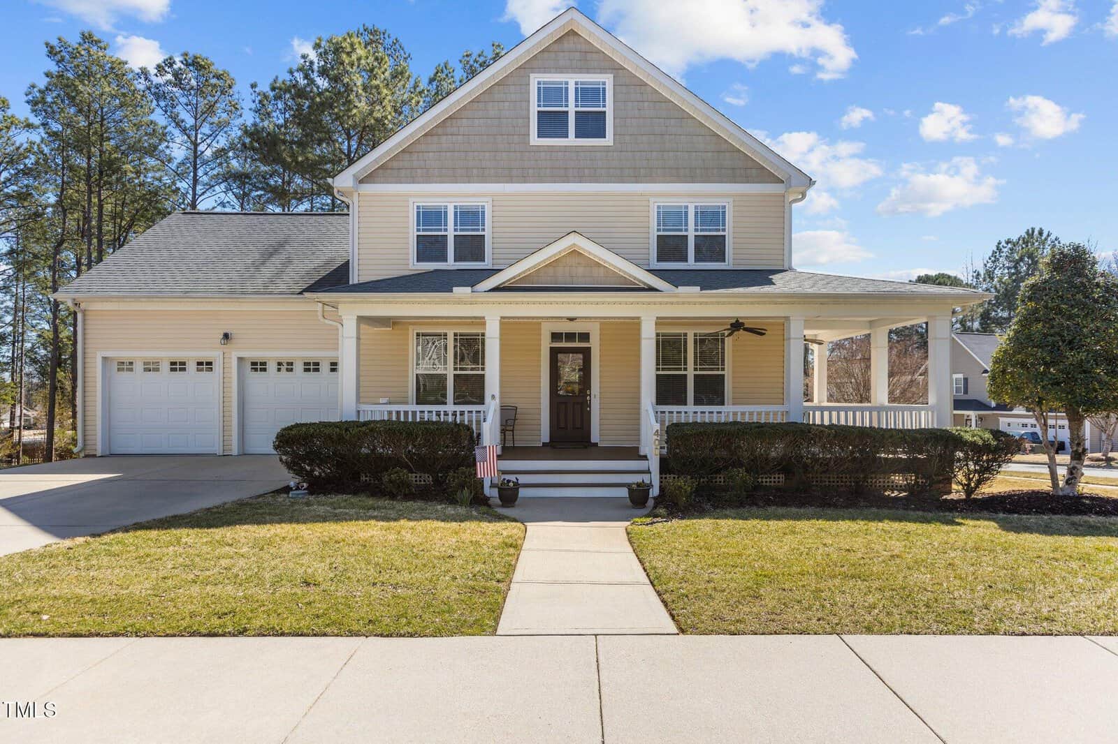 This elegant two-story house at 400 Oxford Park Boulevard boasts beige siding and white trim, featuring a covered front porch and an attached double garage. A manicured lawn and shrubs border the walkway, with trees in the background under a blue sky with clouds.