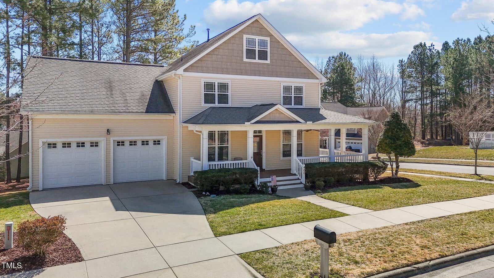 A two-story house with a beige exterior, white trim, and a gabled roof. It has a covered front porch, two-car garage, and a driveway. The lawn is neatly maintained, and there are trees and shrubs around the property. A mailbox is in the foreground.
