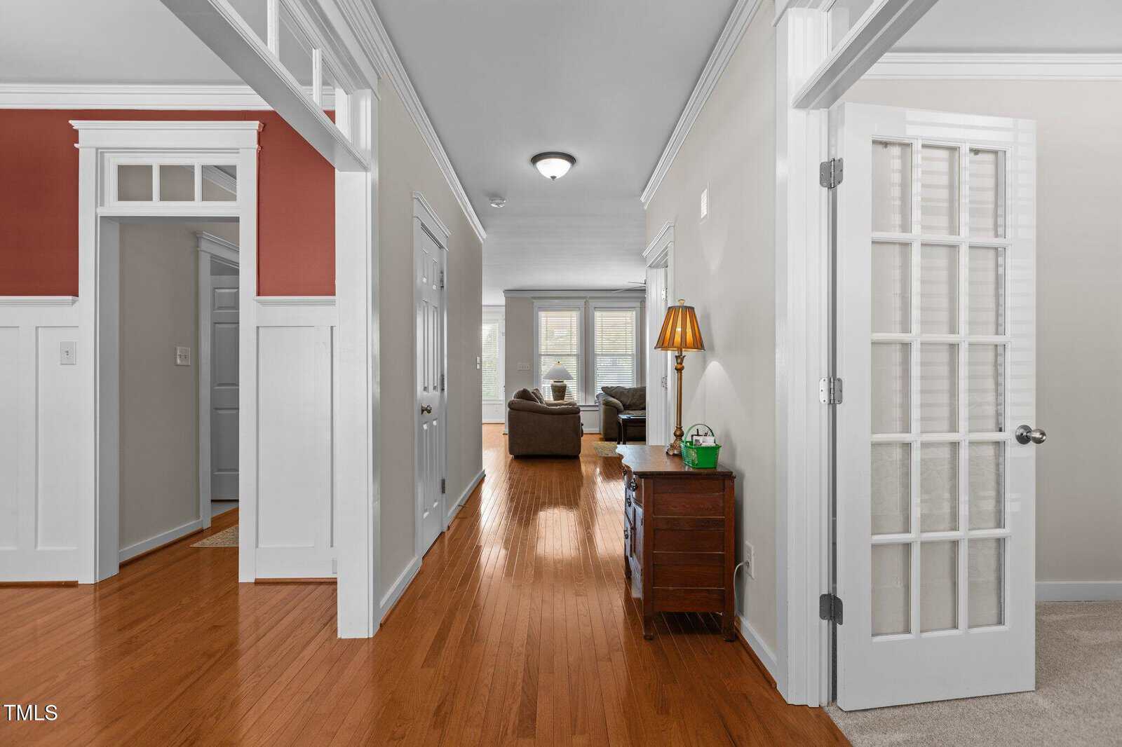 A hallway with wooden floors leads to a sitting area with two couches and a table. The walls are painted white and light beige, with one featuring red accents. A wooden cabinet holds a green lamp, and a room with French doors is on the right.