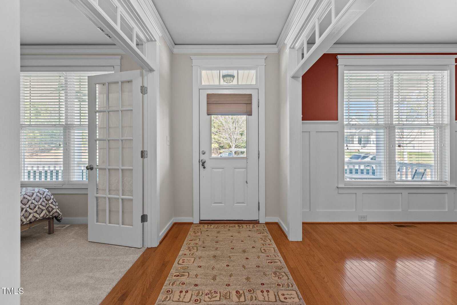 A foyer with a wooden floor and a patterned rug leads to a white front door with a frosted glass window. To the left, a room with a French door and a visible bed. Large windows on each side let in ample light. Walls are painted with white and a deep red accent.