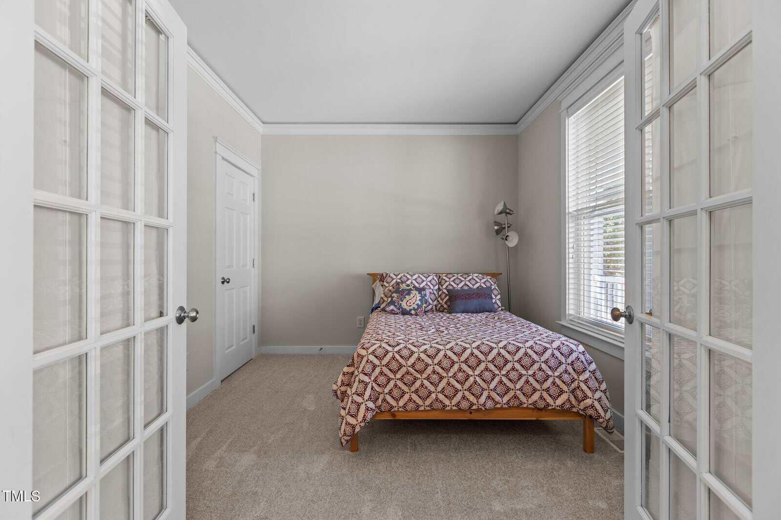 A small bedroom features a wooden bed with a patterned quilt, flanked by two windows with blinds. The room has beige walls and a carpeted floor. Glass-panel double doors open into the space. A closed white door is visible on the left wall.