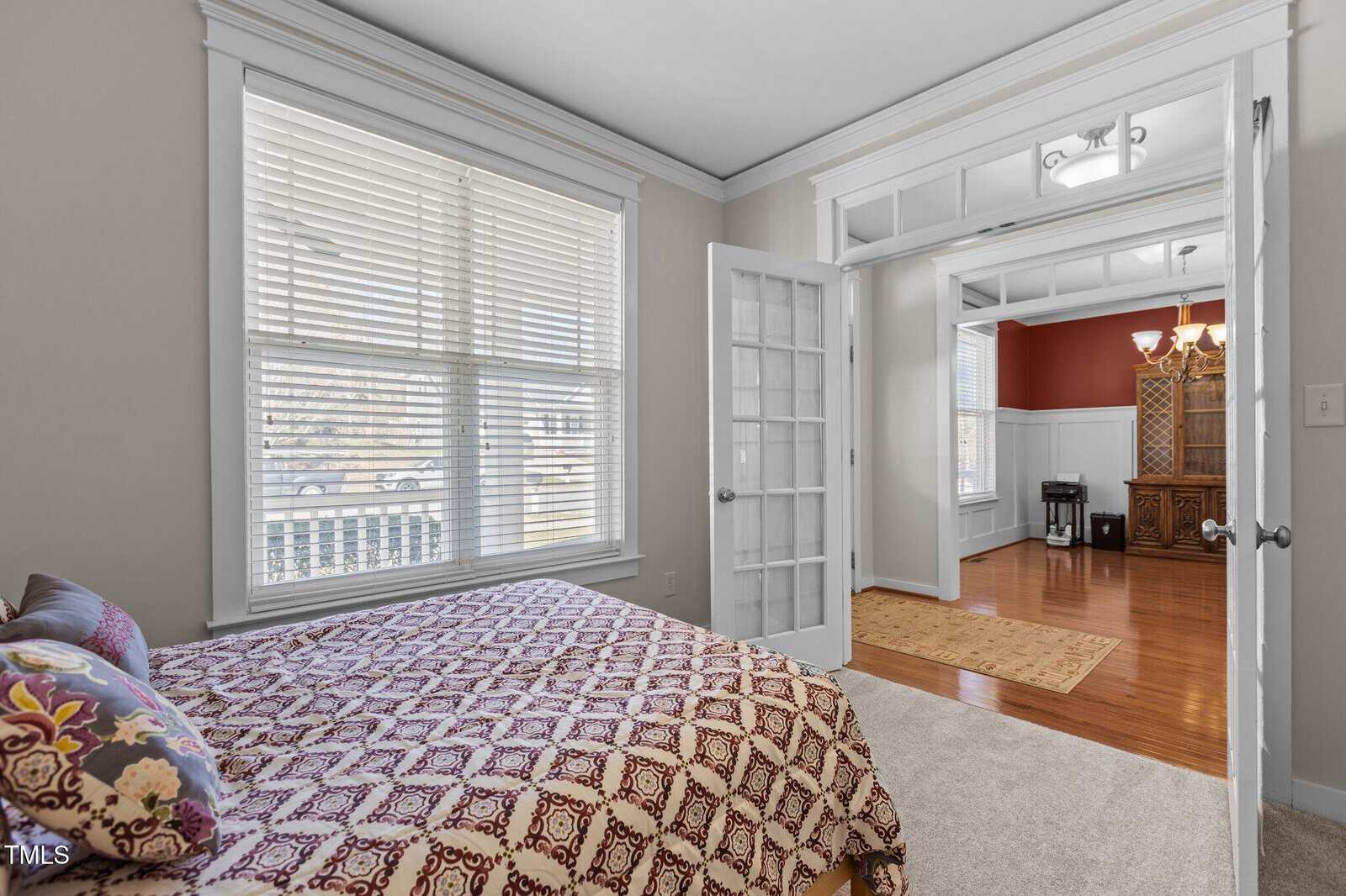 A bedroom with a bed featuring a patterned coverlet and decorative pillows is shown. The room has light-colored walls, a large window with blinds, and an open doorway leading to another room with wood flooring and white paneled walls.