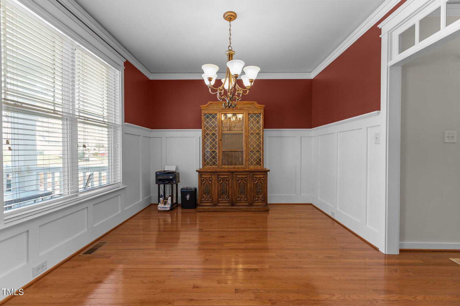 An empty room with white wainscoting and red upper walls. There is a wooden china cabinet, a small printer, and a shredder on the left. The floor is wooden, and a chandelier hangs from the ceiling. Sunlight streams through the large window on the left.